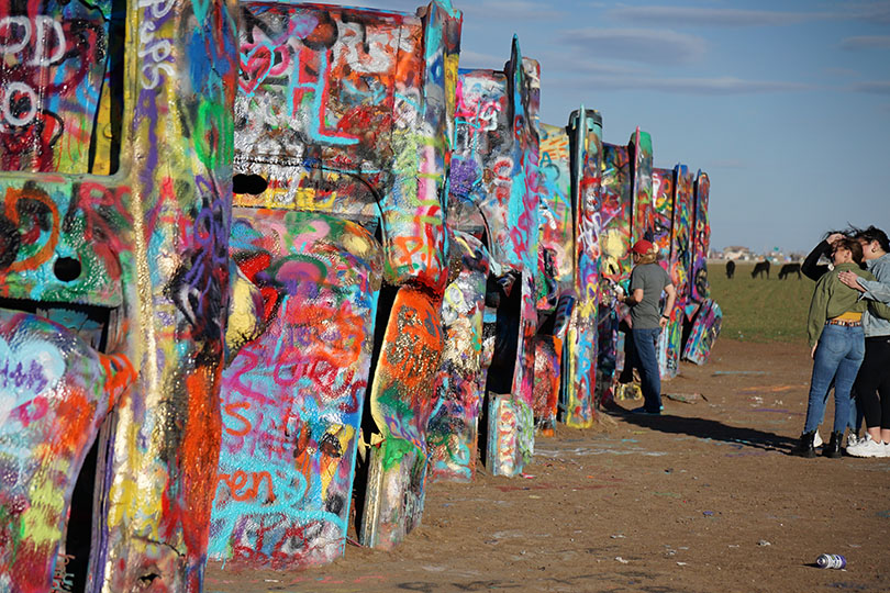 Cadillacs half buried rear end up, covered in spray paint in the middle of a cow field.